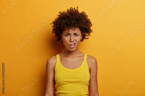 Portrait of dissatisfied dark skinned woman smirks face, purses lips and looks with annoyance at camera, wears casual outfit, discontent hearing something unpleasant, poses indoor over yellow wall