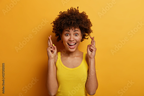 Positive cute girl with Afro hairstyle believes dreams come true, keeps fingers crossed, awaits for something good happened, dressed casually, laughs and looks directly at camera, poses indoor
