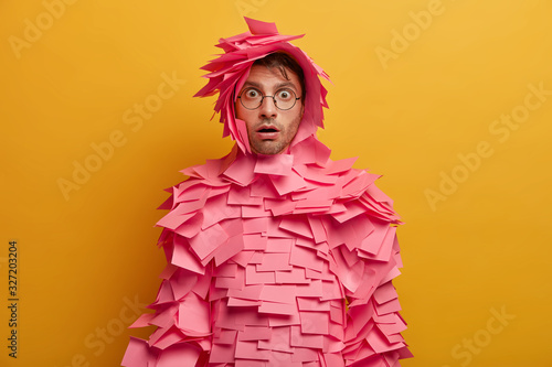 Indoor shot of shocked young Caucasian man stares with bugged eyes, wears transparent glasses, has creative costume made of sticky notes, stares startled and impressed, isolated over yellow wall photo