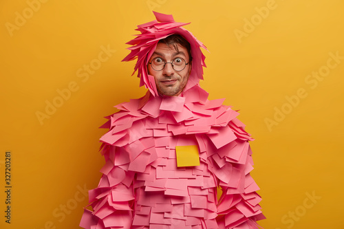 Indoor shot of surprised funny man looks through transparent glasses, wears paper costume has direct gaze at camera isolated over yellow background. Wondered guy dressed in outfit made of sticky notes photo
