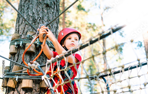 Little boy with climbing gear climbing rope trail between pine trees in adventure park. Boy enjoys climbing in the ropes course adventure. Rope park.