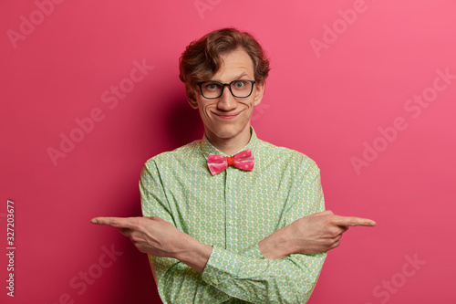 Positive funny man points hands sideways with crossed arms over chest, gives variants, chooses bertween two options, indicates right and left, wears elegant green shirt with bow tie, eyewear photo