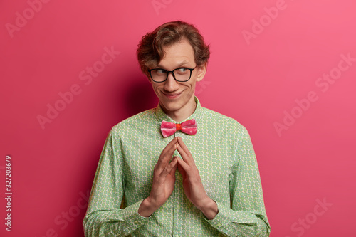 Isolated shot of cunning thoughtful man has evil genius plan, steepls fingers with intention to do something, has pleased pensive expression, wears elegant green shirt and bow tie, stands indoor photo