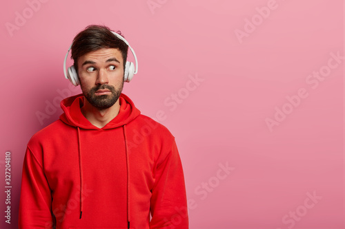 Photo of bearded hipster guy looks away with surprised wondered look, dressed in red sweatshirt, sees something unbelievable, uses headphones, isolated over rosy pastel wall, copy space aside