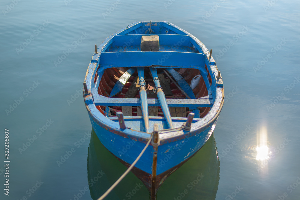Blue Boat in lake. Sunshine on blue water. Wooden blue boat on calm water tied to the harbor by a rope. 