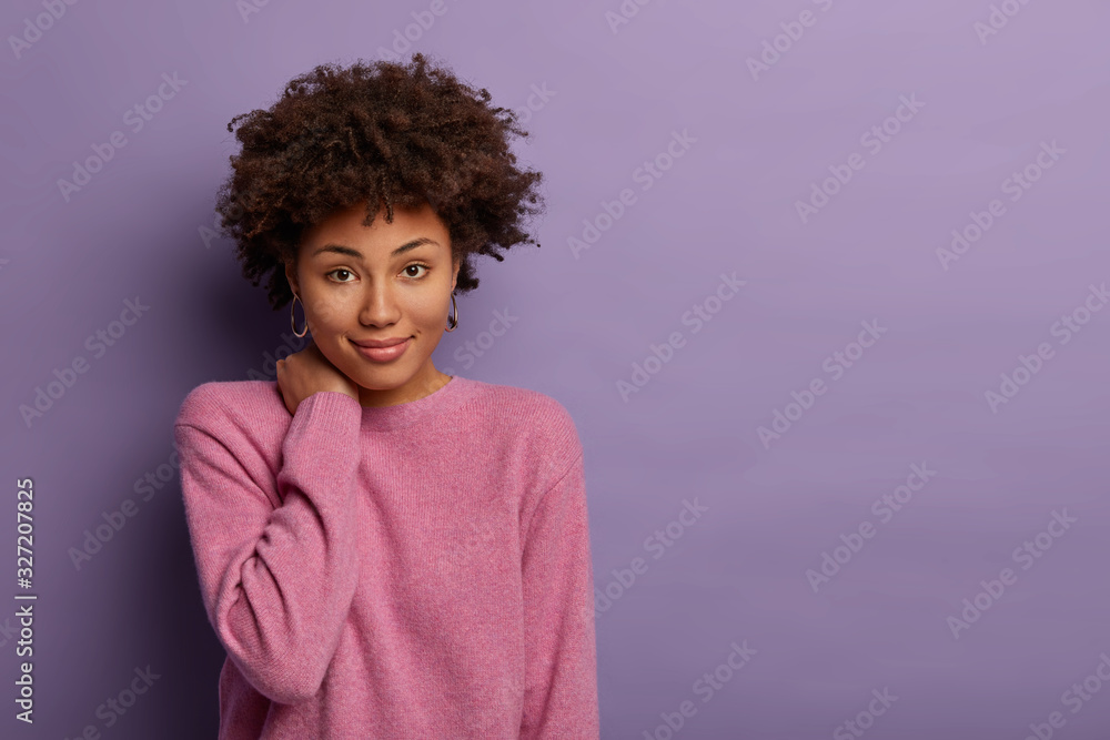 Feminine model with curly hair touches neck, has gentle look, delighted smile, calm expression, casual talk with friend, wears long sleeved jumper, poses over purple background, blank space aside. Feminine model with curly hair touches neck, has gentle look, delighted smile, calm expression, casual talk with friend, wears long sleeved jumper, poses over purple background, blank space aside.