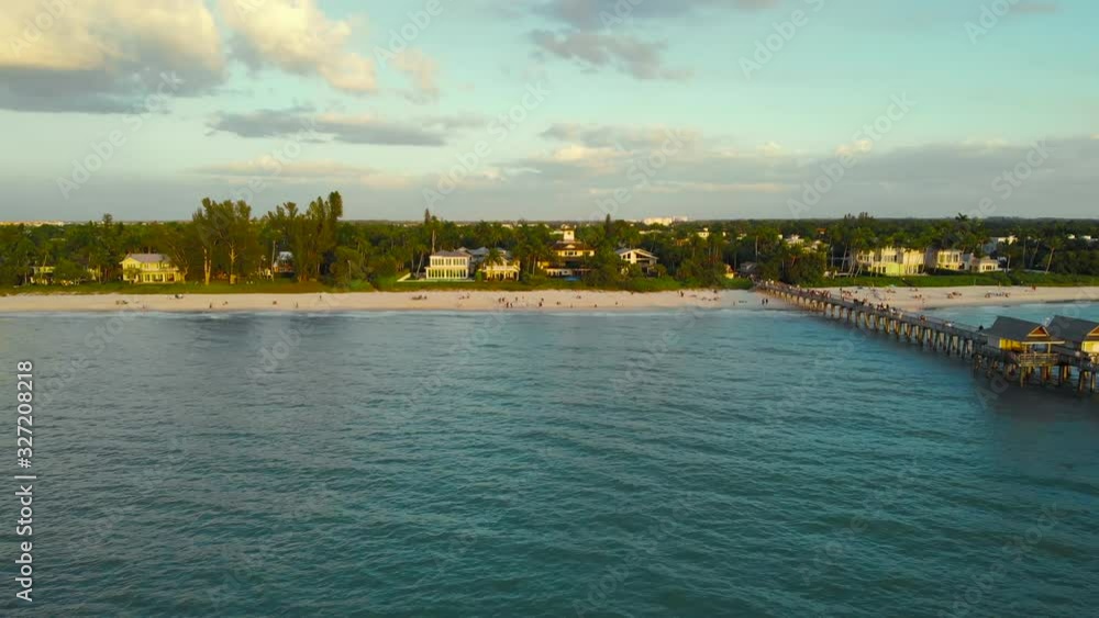 Naples Beach and Fishing Pier at Sunset, Florida. Naples, Florida is a tourist town in the USA. Drone flies forward from the ocean to the coast. People walk along the ocean, aerial view. Naples Beach and Fishing Pier at Sunset, Florida. Naples, Florida is a tourist town in the USA. Drone flies forward from the ocean to the coast. People walk along the ocean, aerial view.