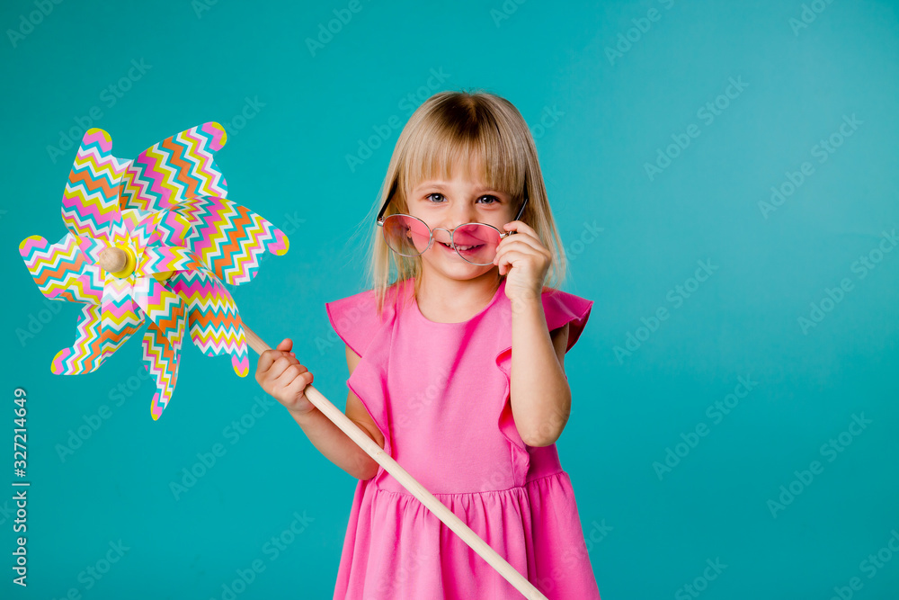child girl blonde smiling in sunglasses in a pink dress holding a windmill on a blue background of isolate. the concept of the summer, space for text child girl blonde smiling in sunglasses in a pink dress holding a windmill on a blue background of isolate. the concept of the summer, space for text
