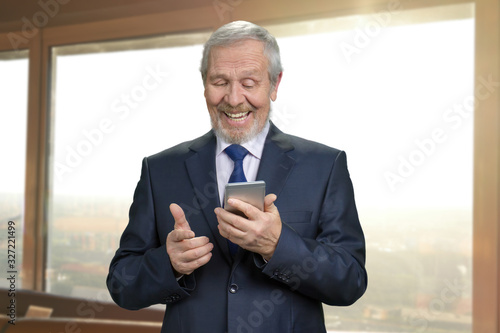 Old man in suit looking at the phone screen. Portrait of smiling senior businessman with gadget. Office windows background.