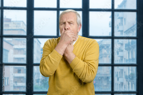Portrait of coughing sick old man. Grandpa in wool yellow sweater is feeling bad against checkered windows background.