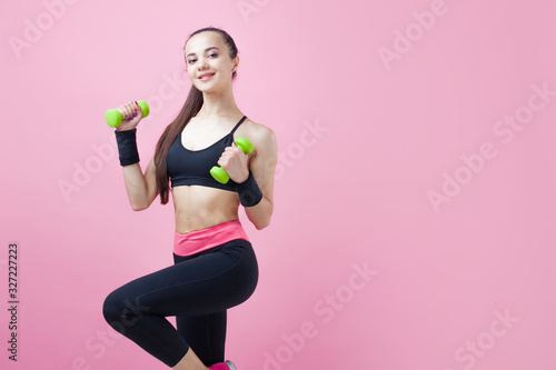 A young athletic woman, with a smile, in black sports top, doing aerobics on pink background with green light dumbbells.