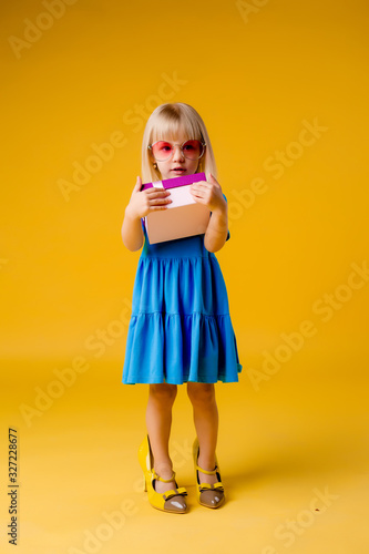 little blonde girl in a blue dress and colored glasses is holding a pink gift box on a yellow background