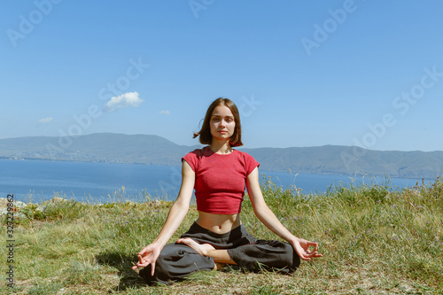 Young woman doing yoga in lotus position and meditating in nature.  Pacified female sitting in asana on grass with lake behind while looking at camera in mountain landscape