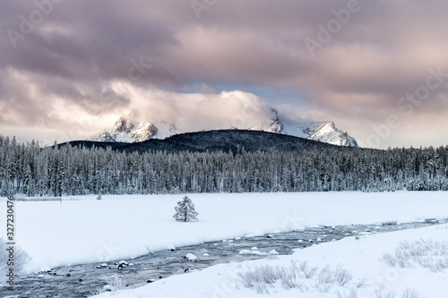 Fringed cold river flows across a snow covered meadow with high mountain peaks
