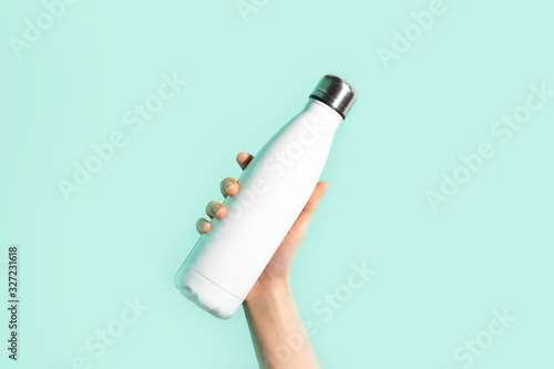 Close-up of female hand, holding white reusable steel stainless eco thermo water bottle, isolated on background of cyan, aqua menthe color. Be plastic free. Zero waste.