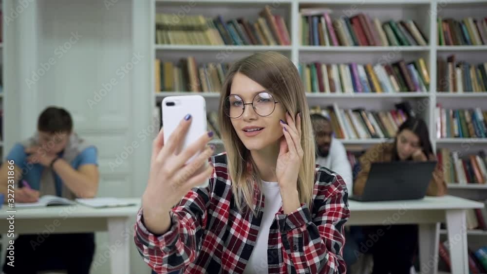 Beautiful smiling modern bright female student in glasses sitting in the library and watching into the phone camera on the others diverse students background Beautiful smiling modern bright female student in glasses sitting in the library and watching into the phone camera on the others diverse students background