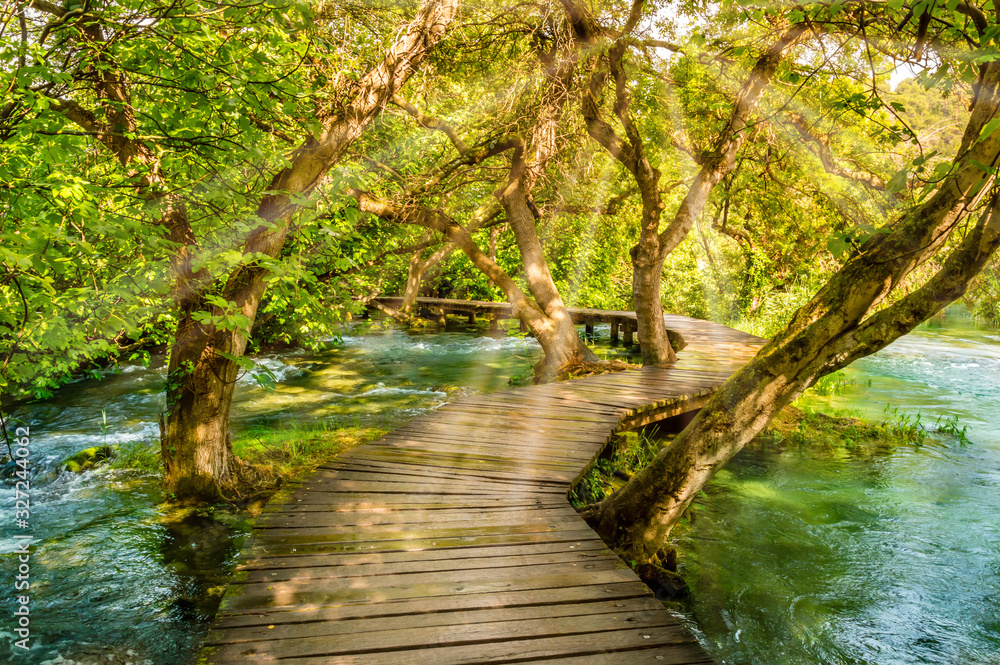 Fototapeta premium Wooden footpath over river in forest of Krka National Park, Croatia. Beautiful scene with trees, water and sunrays.