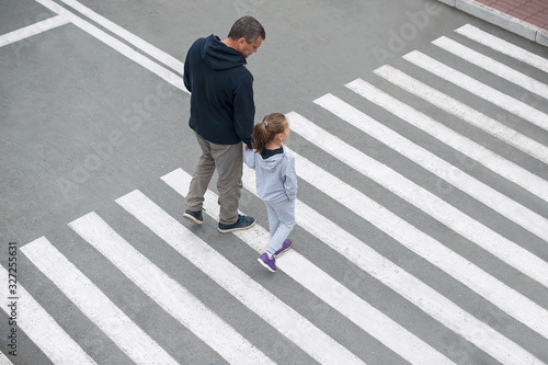 A man and a small child on a zebra crossing trespassing by crossing the street. In the summer on the street kid girl with her father in fashion clothes cross the road. From top view