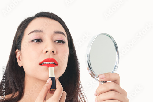 A beautiful Asian woman is carrying a mirror, she is doing makeup and putting red lipstick on her mouth. White background. Beauty concept Body care