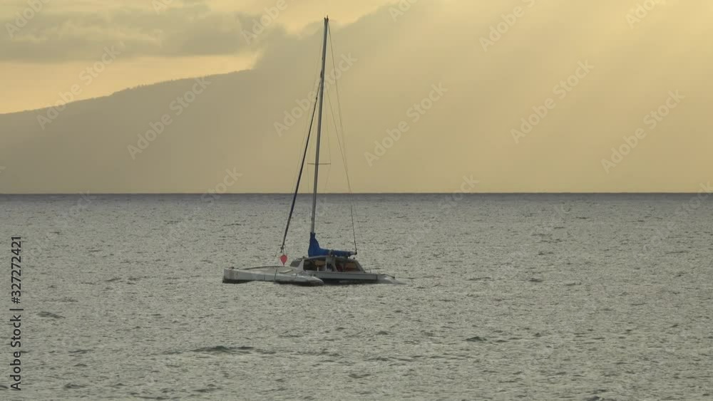 A small, anchored catamaran boat rides the gentle ocean waves in front of a beautiful, golden sky in the early evening. A small, anchored catamaran boat rides the gentle ocean waves in front of a beautiful, golden sky in the early evening.