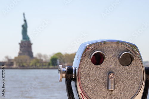 Tourist binoculars on Ellis Island with Statue of Liberty visible in the blurred background. There is a blue sky and water visible. The metal shows age and is burnished