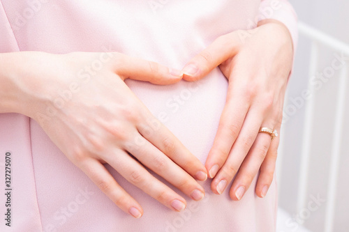 pregnant woman in a dress holds her hands on her stomach in the shape of a heart. The concept of pregnancy, motherhood, preparation and expectation. Close-up indoors.