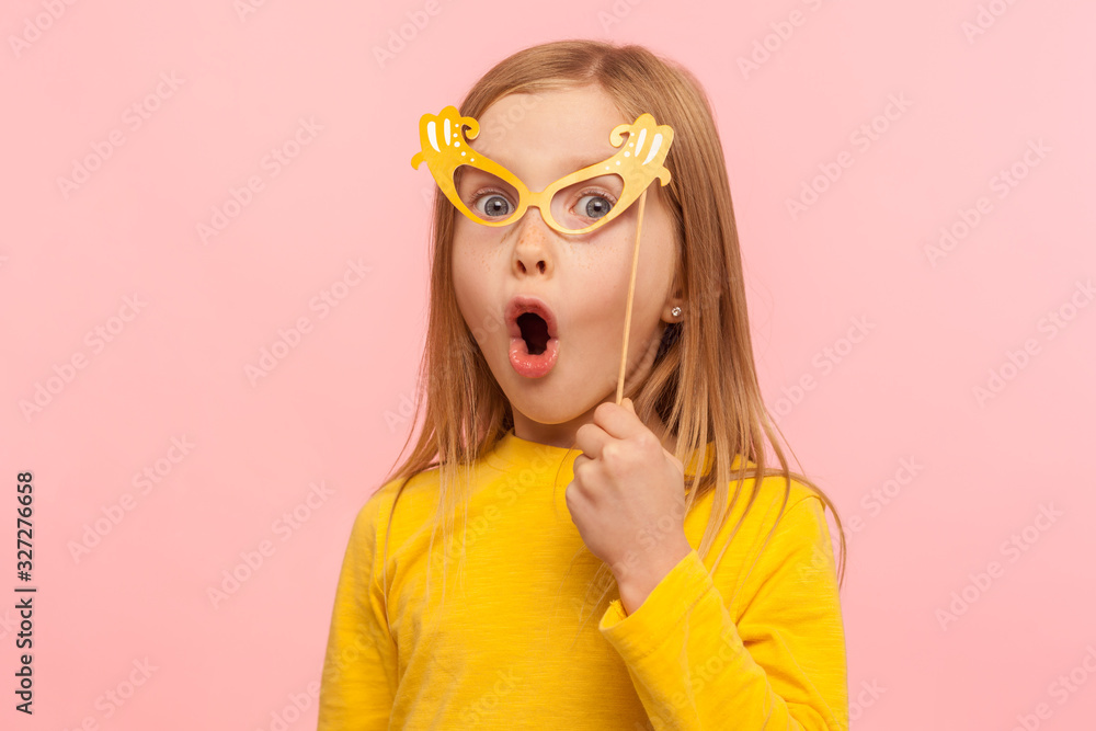 Surprised little girl covering eyes with paper glasses and sticking out tongue, naughty child having fun, wearing masquerade mask and looking shocked. indoor studio shot isolated on pink background Surprised little girl covering eyes with paper glasses and sticking out tongue, naughty child having fun, wearing masquerade mask and looking shocked. indoor studio shot isolated on pink background