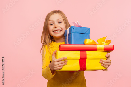Delighted little girl embracing lot of gift boxes and smiling at camera with expression of sincere childish happiness, enjoying perfect birthday with many presents. indoor studio shot, pink background photo