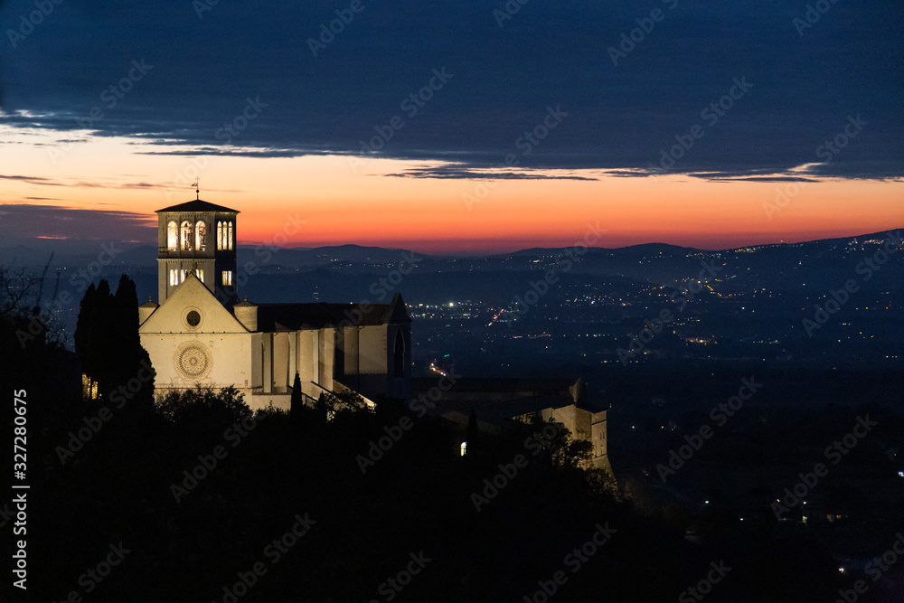 View of St. Francis papal church in Assisi at night, with city lights on the background