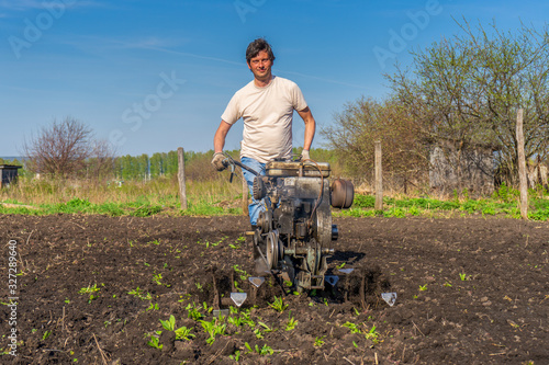 Man in wellingtons with cultivator ploughing ground in sunny day. Farmer plowing kitchen-garden in suburb. Land cultivation, soil tillage. Spring work in garden. Gardening concept