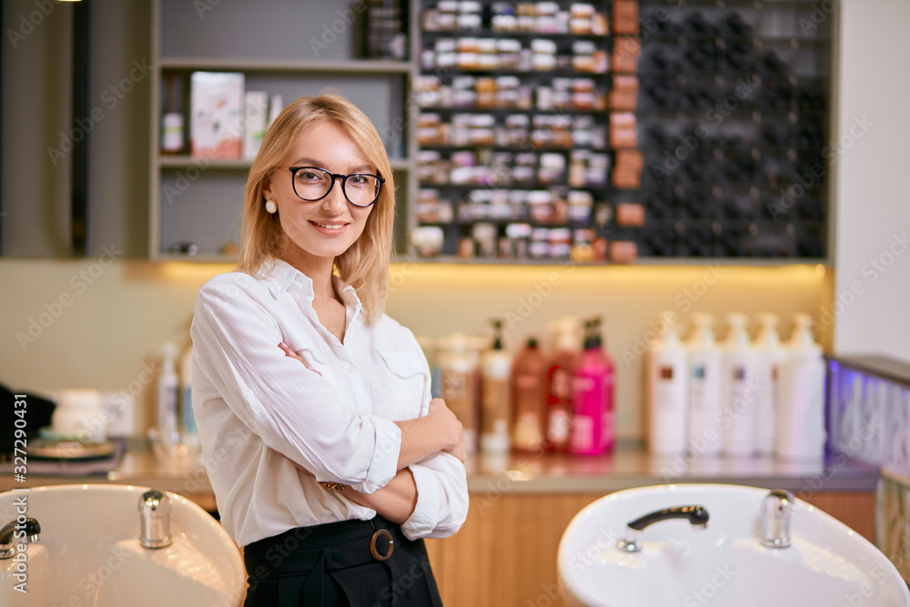 portrait of smiling open-minded consultant of beauty saloon selling beauty products, different shampoos and balms. Blond woman in white shirt and eye glasses stand isolated in beauty saloon portrait of smiling open-minded consultant of beauty saloon selling beauty products, different shampoos and balms. Blond woman in white shirt and eye glasses stand isolated in beauty saloon