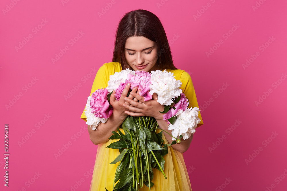 Close up portrait of beautiful brunette woman holding flowers isolated over pink studio background, lady wearing yellow dress embracing big bouquet, girl looking at flowers. 8 march concept.