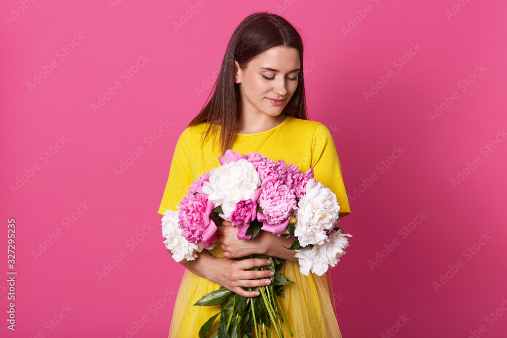 Horizontal shot of gentle woman holding pink and white peony flowers, stands smiling isoalted over rosy background, brunette lady wearing yellow dress, having present for 8 march. Women's day. Horizontal shot of gentle woman holding pink and white peony flowers, stands smiling isoalted over rosy background, brunette lady wearing yellow dress, having present for 8 march. Women's day.