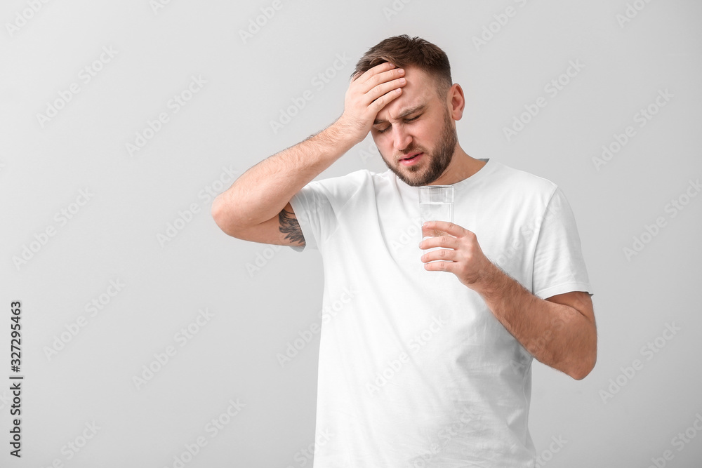 Young man with glass of water suffering from headache on light background