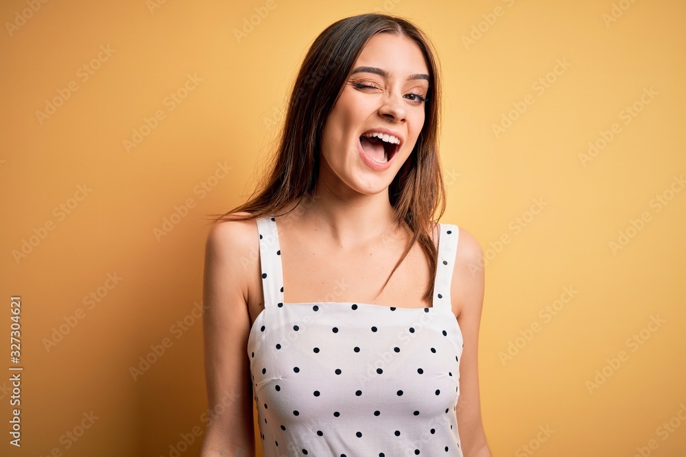 Young beautiful brunette woman wearing casual dress standing over yellow background winking looking at the camera with sexy expression, cheerful and happy face. Young beautiful brunette woman wearing casual dress standing over yellow background winking looking at the camera with sexy expression, cheerful and happy face.