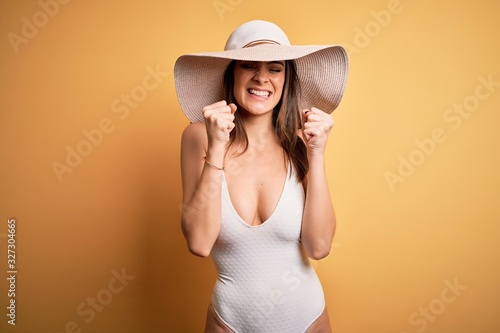 Young beautiful brunette woman on vacation wearing swimsuit and summer hat excited for success with arms raised and eyes closed celebrating victory smiling. Winner concept.