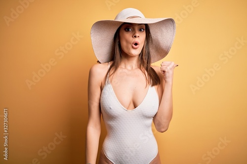 Young beautiful brunette woman on vacation wearing swimsuit and summer hat Surprised pointing with hand finger to the side, open mouth amazed expression.