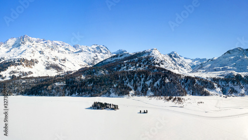 Aerial image of the Chaviolas islets and Isola with people doing sports on the snow-covered frozen lake surface of Silsersee, St. Moritz, Switzerland