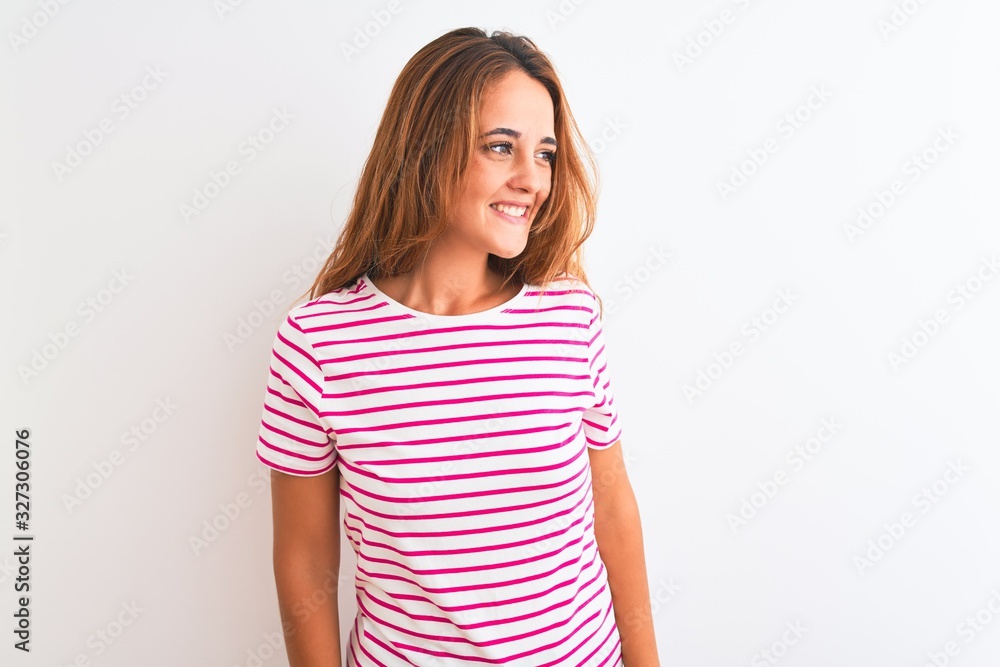 Young redhead woman wearing striped casual t-shirt stading over white isolated background looking away to side with smile on face, natural expression. Laughing confident. Young redhead woman wearing striped casual t-shirt stading over white isolated background looking away to side with smile on face, natural expression. Laughing confident.