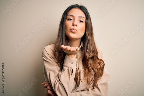 Young beautiful brunette woman wearing casual shirt standing over white background looking at the camera blowing a kiss with hand on air being lovely and sexy. Love expression.