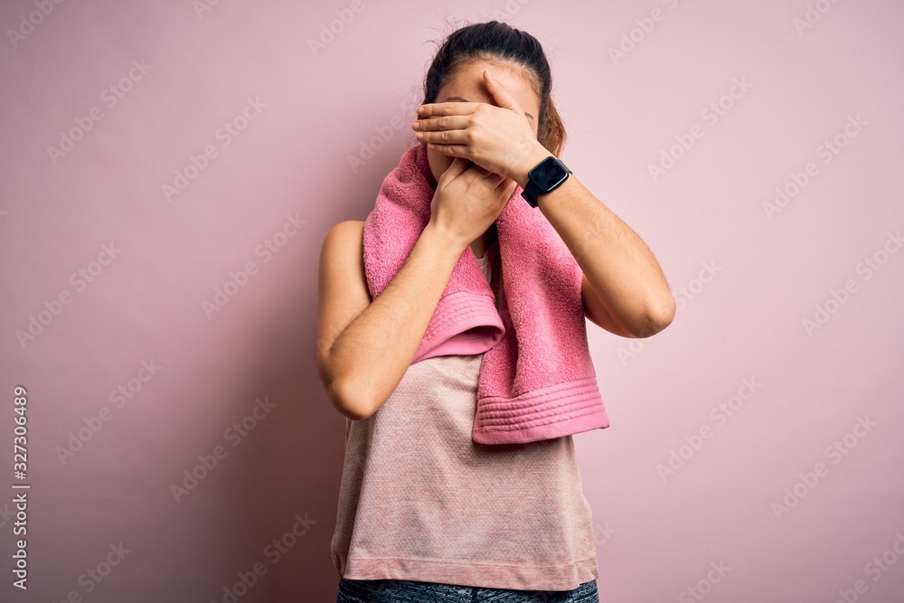 Young beautiful brunette sportswoman wearing sportswear and towel over pink background Covering eyes and mouth with hands, surprised and shocked. Hiding emotion Young beautiful brunette sportswoman wearing sportswear and towel over pink background Covering eyes and mouth with hands, surprised and shocked. Hiding emotion