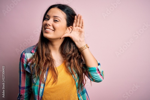 Young beautiful brunette woman wearing casual colorful shirt standing over pink background smiling with hand over ear listening an hearing to rumor or gossip. Deafness concept.