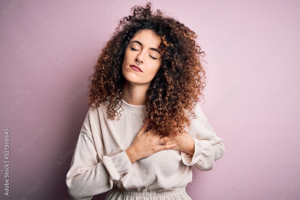 Beautiful woman with curly hair and piercing wearing casual sweater over pink background smiling with hands on chest with closed eyes and grateful gesture on face. Health concept. Beautiful woman with curly hair and piercing wearing casual sweater over pink background smiling with hands on chest with closed eyes and grateful gesture on face. Health concept.