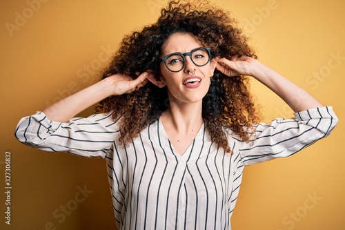 Young beautiful woman with curly hair and piercing wearing striped shirt and glasses Smiling pulling ears with fingers, funny gesture. Audition problem