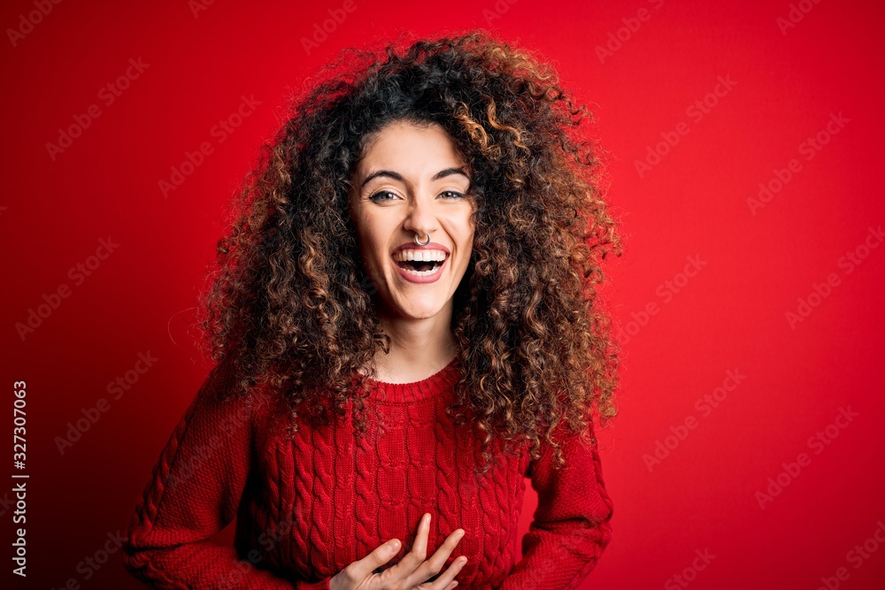 Young beautiful woman with curly hair and piercing wearing casual red sweater smiling and laughing hard out loud because funny crazy joke with hands on body. Young beautiful woman with curly hair and piercing wearing casual red sweater smiling and laughing hard out loud because funny crazy joke with hands on body.