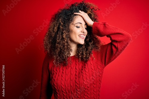 Young beautiful woman with curly hair and piercing wearing casual red sweater smiling confident touching hair with hand up gesture  posing attractive and fashionable
