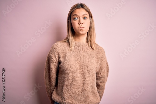 Young beautiful blonde woman wearing winter wool sweater over pink isolated background puffing cheeks with funny face. Mouth inflated with air, crazy expression. © Krakenimages.com