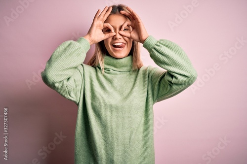 Young beautiful blonde woman wearing winter wool sweater over pink isolated background doing ok gesture like binoculars sticking tongue out, eyes looking through fingers. Crazy expression. © Krakenimages.com