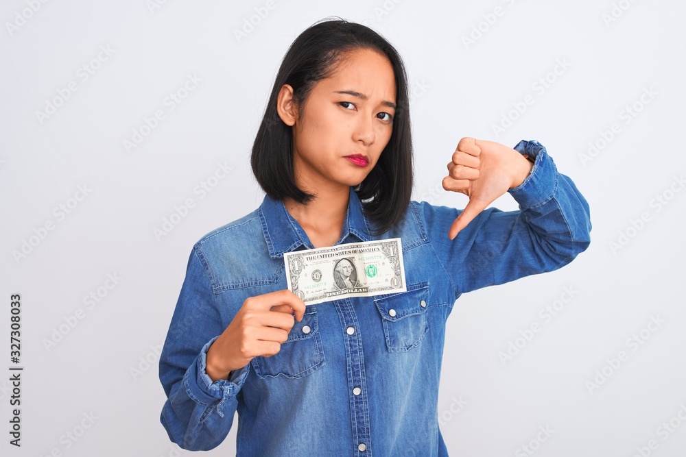 Young beautiful chinese woman holding one dollar standing over isolated white background with angry face, negative sign showing dislike with thumbs down, rejection concept Young beautiful chinese woman holding one dollar standing over isolated white background with angry face, negative sign showing dislike with thumbs down, rejection concept