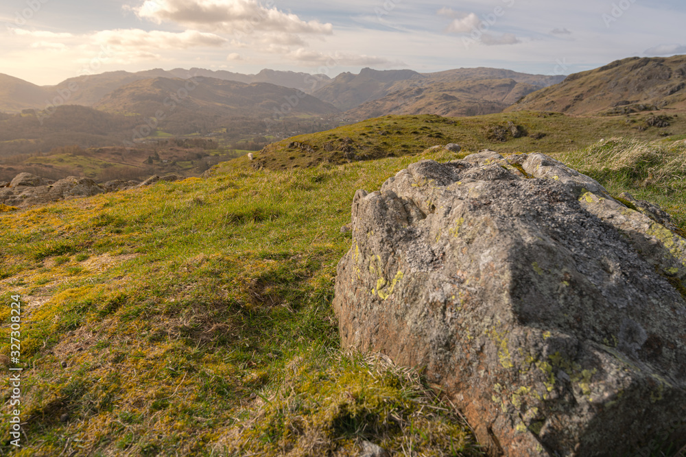 Fields, hills, mountains and a large rock in the foreground at the Lake District National Park, UK during sunset